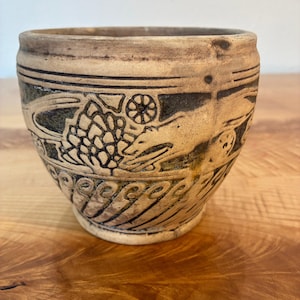 May include: A beige ceramic bowl with a dark brown incised design featuring a stylized animal, a pinecone-like shape, and circular motifs. The lower section of the bowl is decorated with a repeating pattern of curved, wave-like lines.