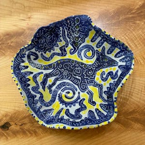 May include: A hand-painted ceramic bowl with an irregular, wavy rim. The interior features a dark blue fox-like creature surrounded by intricate white line patterns, spirals, and sun motifs set against a bright yellow and white background.