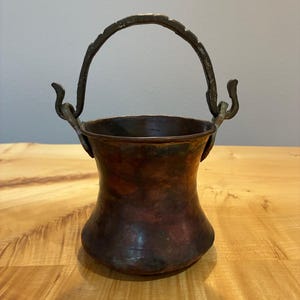 May include: A small, hammered copper pot with a dark, weathered patina and a flared rim. It features a curved, textured metal handle attached to two decorative, upward-curving side brackets. The body of the pot has a rounded, concave shape.