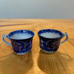 May include: Two small porcelain cups featuring a deep blue and white floral pattern with gold accents. Each cup has a curved handle and a flared rim, showcasing intricate blue detailing around the top edge and a central gold floral motif.