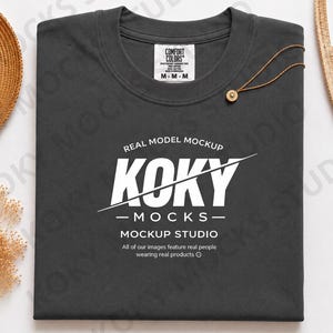 Może przedstawiać: Ciemnoszary t-shirt z okrągłym dekoltem i białym nadrukiem graficznym o treści REAL MODEL MOCKUP KOKY MOCKS MOCKUP STUDIO. Koszulka posiada metkę marki Comfort Colors przy wewnętrznym dekolcie oraz mały złoty naszyjnik z zawieszką.