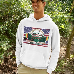 May include: A white hooded sweatshirt featuring a graphic print of the American flag, a helicopter, a green clover, and the text 4th Infantry Division, Not For Fame Or Glory But For The Brother Next To Me, Steadfast And Loyal, Vietnam Veteran.