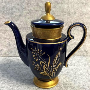 Könnte beinhalten: Eine tief kobaltblaue Keramik-Teekanne mit einem goldfarbenen Blumenmuster auf dem Körper. Die Teekanne verfügt über einen passenden Deckel mit goldenem Knauf, einen goldfarbenen, texturierten Fuß, einen texturierten Kragen sowie einen goldenen Ausguss und Henkel.