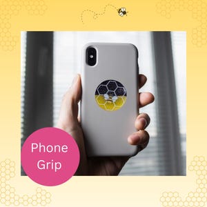 May include: A light gray smartphone case featuring a circular phone grip. The grip displays a graphic of a bee centered over a honeycomb pattern, with the top half in black with white speckles and the bottom half in a solid yellow color.