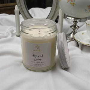 May include: A glass jar candle with a white lid, labeled "Royal Linens." The candle has a creamy white wax and a visible wick. The label includes the scent description "Ozone, Linen, Musk." The candle is handcrafted.