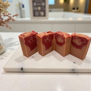 May include: Four bars of handmade soap with a layered design of red and beige hues. The soap is displayed on a white marble tray. The soap bars have the text "VANITY SOAPS" printed on the side.