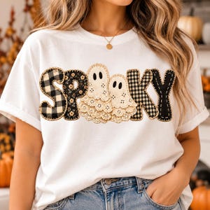 Może przedstawiać: Biały t-shirt z krótkim rękawem z napisem SPOOKY na piersi. Litery są ozdobione czarno-białą kratą i motywami kwiatowymi, a dwie kremowe koronkowe duszki zastępują literę O.