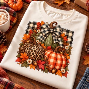 Peut inclure: Un sweat-shirt crème arborant un motif de trois citrouilles décorées d'imprimés léopard, à carreaux orange et verts. Le design comprend un nœud à carreaux noir et blanc, des feuilles d'automne, des glands et de petites fleurs blanches.