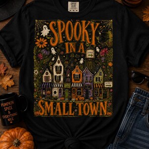 Peut inclure: Un t-shirt noir avec une impression graphique représentant une petite ville aux bâtiments colorés, des fantômes et des citrouilles. Le texte "SPOOKY IN A SMALL TOWN" est écrit en orange. Le motif inclut des fleurs, un château d'eau et des panneaux.