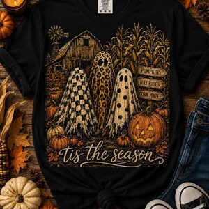 Peut inclure: Un t-shirt noir avec un motif représentant trois fantômes aux motifs damier, léopard et pois devant une grange et un champ de maïs. Le design inclut une citrouille d'Halloween, des panneaux en bois indiquant PUMPKINS, HAY RIDES, CORN MAZE et le texte tis the season.
