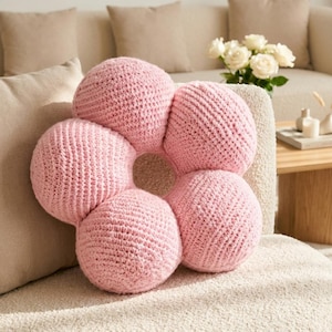 Könnte beinhalten: Ein hellrosa, blumenförmiges Dekokissen mit einer texturierten Häkelstrickoptik. Das Kissen besteht aus fünf abgerundeten, gefüllten Blütenblättern, die kreisförmig um eine hohle, offene Mitte angeordnet sind und ein weiches, plüschiges Aussehen haben.