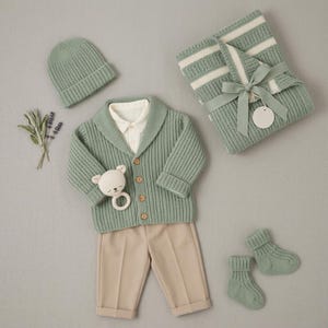 Könnte beinhalten: Eine salbeigrüne, gerippte Strickjacke, eine passende Mütze und Socken, kombiniert mit einer beigen Hose und einem weißen Hemd mit Kragen. Dazu eine gefaltete, salbeigrün-weiß gestreifte Decke mit Schleife und eine bärenförmige Rassel.