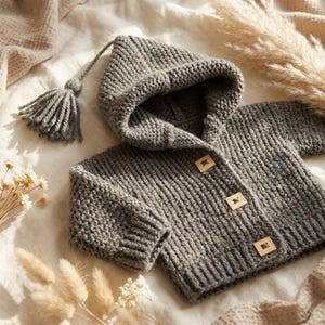 Könnte beinhalten: Eine anthrazitfarbene Strickjacke mit Kapuze, gerippter Textur, langen Ärmeln und drei quadratischen Holzknöpfen auf der Vorderseite. Eine passende Garnquaste ist mit einer langen, dünnen geflochtenen Kordel an der Kapuze befestigt.