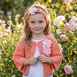 Könnte beinhalten: Eine korallenfarbene Häkeljacke mit Dreiviertelärmeln und passendem Stirnband. Die Jacke ist mit einer vertikalen Reihe aus drei strukturierten, mehrfarbigen Blumenapplikationen auf der linken Seite versehen, das Stirnband hat einen Blumenakzent.