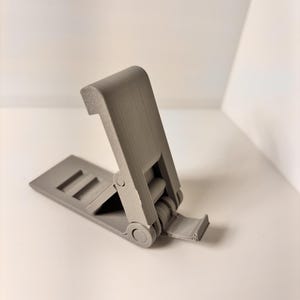 May include: A gray, hinged plastic phone stand, open and angled. The stand has a rectangular base with two parallel slots and a hinged top section. The stand is designed to hold a phone upright.