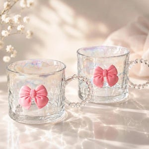 Könnte beinhalten: Zwei transparente Glasbecher mit schillernden Oberflächen und strukturierten Oberflächen. Jeder Becher hat eine rosa Schleife und einen Griff, der mit kleinen, klaren Perlen verziert ist. Die Becher stehen auf einer reflektierenden Oberfläche.