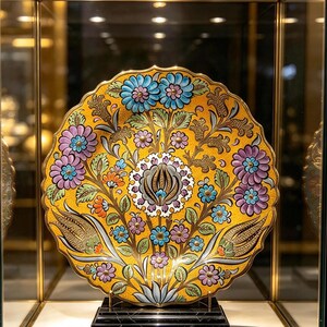 May include: A decorative ceramic plate with a scalloped edge, featuring a vibrant yellow background adorned with intricate floral patterns in shades of blue, purple, and orange, displayed on a black marble base within a glass enclosure.