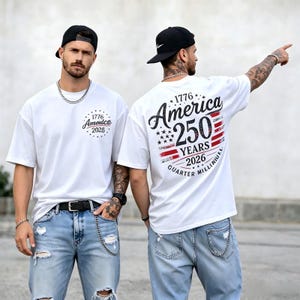 Könnte beinhalten: Ein weißes Kurzarm-T-Shirt mit einer kleinen Grafik auf der linken Brust und einer großen Grafik auf der Rückseite. Das Design enthält den Text 1776 America 250 Years 2026 Quarter Millinniel, Sterne und ein rot-weiß-blaues Flaggenmotiv.