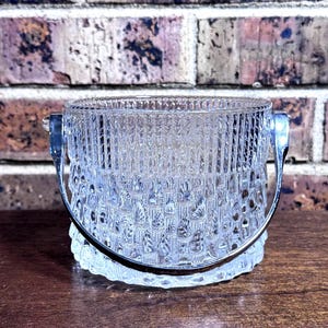 May include: A clear glass ice bucket featuring a textured, geometric pattern on the exterior surface. The cylindrical container is equipped with a polished silver-toned metal swing handle attached to two side mounts, providing a functional grip.