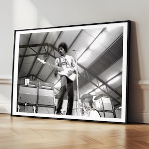 May include: Black and white framed photograph of a musician playing an electric guitar on stage. The image features amplifiers, drums, and a microphone stand. The musician is wearing a patterned shirt and dark trousers.