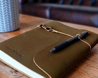 Puede incluir: Un diario de cuero verde oliva con cubierta envolvente, que presenta un cierre de metal color latón y un bolígrafo retráctil negro asegurado en una trabilla de cuero. La cubierta tiene grabado el texto Crafty Horse y un icono de caballo.