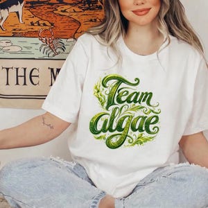 Peut inclure: Un t-shirt blanc à manches courtes avec une impression graphique sur la poitrine portant le texte Team Algae dans une police verte stylisée et tourbillonnante. Le motif comprend des détails complexes de feuilles et de vignes autour du texte.
