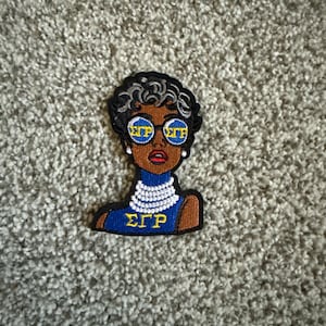 May include: An embroidered patch featuring a stylised figure with short dark hair, wearing round blue sunglasses with the Greek letters ΣΓΡ in yellow. The figure wears a white multi-strand necklace and a blue top with the yellow Greek letters ΣΓΡ.