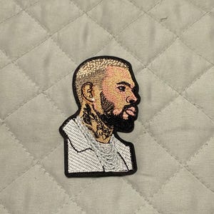 May include: Embroidered patch featuring a side profile of a man with a beard, tattoos, and a chain. The patch has a black border and is set against a quilted background. The man's hair is light-colored, and his skin is a mix of pink and brown.