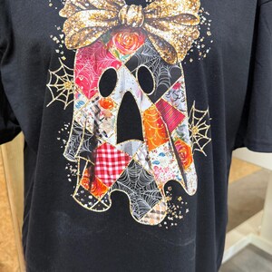 Könnte beinhalten: Ein schwarzes T-Shirt mit einer großen Geistergrafik. Der Geist ist mit einem Patchwork aus bunten Blumen-, Karomustern und Spinnennetzen gefüllt. Eine große, glitzernde goldene Schleife sitzt auf dem Kopf des Geistes, umgeben von Goldglitzer.