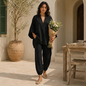May include: A black two-piece set consisting of a loose-fitting V-neck tunic with wide sleeves and matching tapered trousers with elasticated cuffs. The outfit is worn with simple beige flat shoes while holding a bouquet of white flowers.