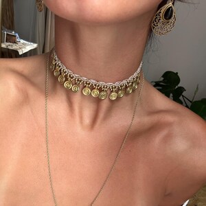 Op de afbeelding: Een witte kanten choker-halsketting met een rij kleine, goudkleurige metalen bedels met spiraalpatronen. De ketting wordt gecombineerd met een lange, dunne goudkleurige ketting en bijpassende druppelvormige filigraan oorbellen met zwarte stenen.