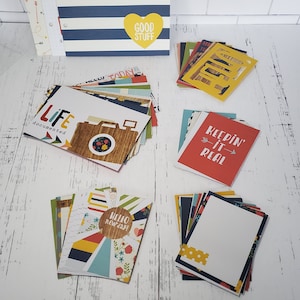 May include: Assortment of colorful paper crafting supplies including cardstock, patterned paper, and die cuts. The paper features various designs and phrases like "Life documented", "Hello New Day!" and "Keepin' it Real".