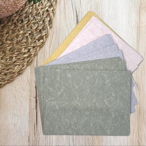 May include: A stack of green, gray, pink, and yellow paper envelopes. The envelopes are arranged in a fan shape, with the green envelopes in the foreground. The paper has a textured appearance. The background is a light wood surface.