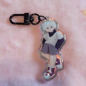 May include: An acrylic keychain featuring a glittery, anime-style character with spiky white hair, wearing a white short-sleeved shirt, dark purple long sleeves, purple shorts, and purple and white sneakers, attached to a black metal lobster clasp.