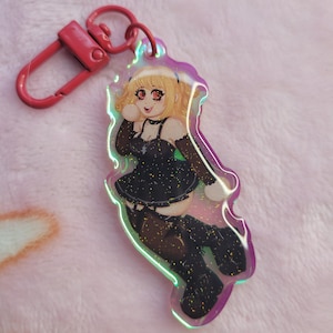 May include: A clear acrylic keychain featuring a glittery illustration of a blonde character with red eyes wearing a black dress, black stockings, and boots. The keychain has a pink metal lobster clasp and a holographic edge that reflects light.