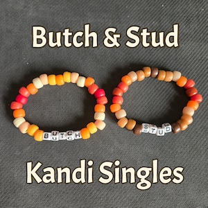 Peut inclure: Deux bracelets élastiques composés de perles en plastique rondes dans des tons orange, rouge et marron. Un bracelet comporte des perles lettres blanches formant le mot BUTCH, l'autre le mot STUD. Le texte Butch & Stud Kandi Singles est inscrit au-dessus et en dessous des bracelets.