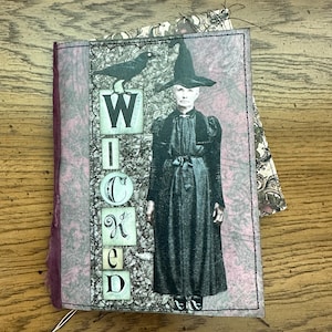 May include: A rectangular journal with a pink and gray textured cover featuring a black and white illustration of a person in a pointed hat and a crow. The word WICKED is printed vertically in black letters on white squares along the left side.
