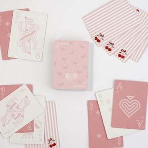 May include: A deck of playing cards with a pink and white color scheme. The cards feature a variety of designs, including cherries, bows, and hearts. The Joker card is visible, along with other cards.