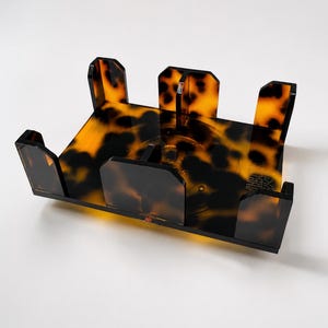 May include: A rectangular acrylic holder featuring a brown and black tortoiseshell pattern. The organizer has a flat base with four vertical side panels and a central divider, designed to hold items in a structured, upright position.