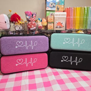 May include: Four rectangular cases in purple, teal, pink, and black, each with a white heart and heartbeat line design. The cases have a zipper closure and a small handle. The cases are on a pink and white checkered surface.