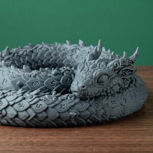 May include: A coiled, grey dragon figurine with detailed scales and rune markings. The dragon's head is resting, with detailed horns and facial features. The sculpture is on a wooden surface against a green backdrop.