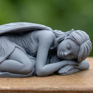 May include: A grey fairy ornament in a sleeping position. The fairy is lying down with its head resting on its arms. It has large, detailed wings and is wearing a dress with leaf-like details. The ornament is on a wooden surface.