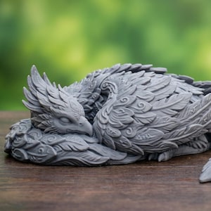 May include: A gray, detailed sculpture of a sleeping mythical bird with feathered wings and tail. The bird is curled up, with its head resting on its body. The sculpture is on a wooden surface with a blurred green background.