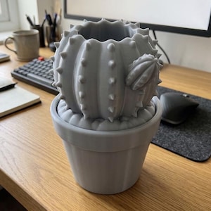 May include: A light gray, 3D-printed cactus-shaped planter featuring a ribbed, spherical body with numerous small, pointed spikes and a smaller bud attached to the side, all resting within a matching, simple, tapered cylindrical plant pot.
