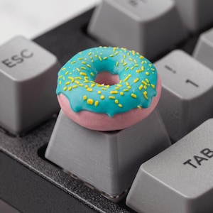 Peut inclure: Une touche de clavier mécanique personnalisée représentant un beignet rose avec un glaçage bleu vif et de petites pépites jaunes. Le beignet est monté sur une touche grise standard située entre les touches ESC et TAB d'un clavier noir.