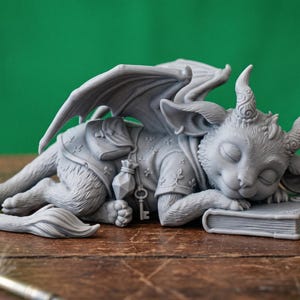 May include: A gray fantasy figurine of a sleeping dragon-like creature with closed eyes, resting on a book. The figurine has wings, horns, and a key pendant. The background is a solid green.