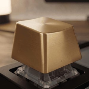 May include: A single gold-colored metallic keycap with a brushed finish and a slightly concave top surface. The keycap is mounted onto a clear plastic mechanical keyboard switch housing, which is set into a black keyboard base frame.