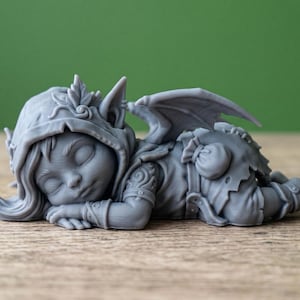 Puede incluir: Una figura gris de una hada durmiente con una capucha, alas y ropa detallada. El hada está acostada con los ojos cerrados, apoyando la cabeza en sus brazos. La figura está sobre una superficie de madera.