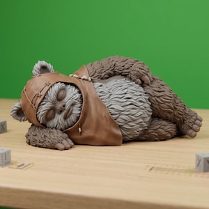 May include: A small, sculpted figurine of a furry creature with grey fur on its chest and brown fur on its limbs, wearing a brown hood with stitched details. The creature is depicted in a sleeping position with its eyes closed and arms tucked in.