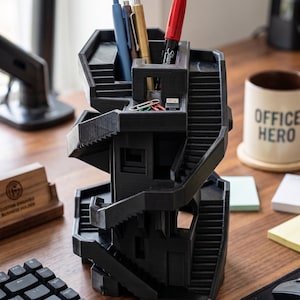 Puede incluir: Un organizador de escritorio negro impreso en 3D con forma de estructura arquitectónica de varios niveles con escaleras interconectadas. Cuenta con múltiples compartimentos para guardar bolígrafos, lápices y clips de papel.